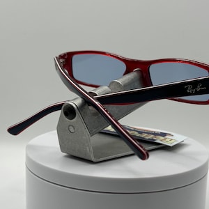 May include: Red and black plastic framed glasses with blue lenses. The glasses are on a silver display stand. The Ray-Ban logo is visible on the right temple.