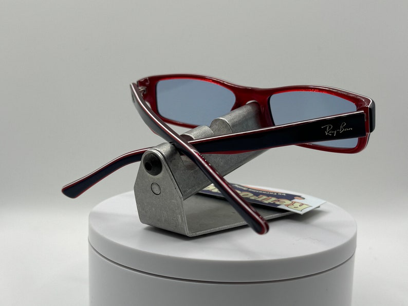May include: Red and black plastic framed glasses with blue lenses. The glasses are on a silver display stand. The Ray-Ban logo is visible on the right temple.