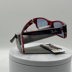 May include: Red and black Ray-Ban sunglasses with blue lenses. The sunglasses are on a silver stand.
