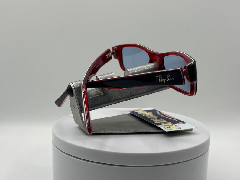 May include: Red and black Ray-Ban sunglasses with blue lenses. The sunglasses are on a silver stand.