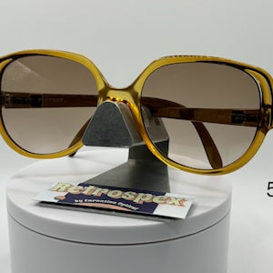 May include: A pair of amber-colored plastic sunglasses with brown lenses. The sunglasses are on a white display stand with a black and white label that reads "Retrospec by Carnation Optical".