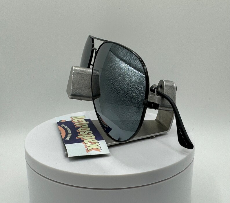 May include: Black aviator sunglasses with mirrored lenses on a silver display stand. The sunglasses have a black frame and are made in China.