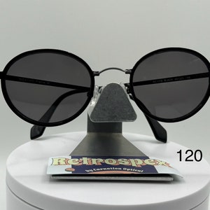 May include: Black round sunglasses with a thin metal frame. The sunglasses are sitting on a white display stand with a sticker that says "Retrospec by Carnation Optical" and the number "120".