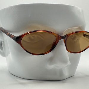 May include: Oval-shaped sunglasses with a tortoiseshell frame and brown lenses. The sunglasses are displayed on a white ceramic head sculpture. The frame has a classic, retro design.