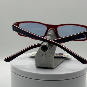 May include: Red and black Ray-Ban sunglasses with blue lenses, folded and displayed on a silver stand.