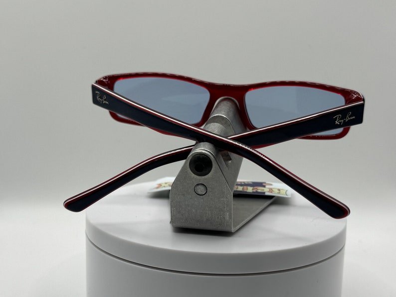 May include: Red and black Ray-Ban sunglasses with blue lenses, folded and displayed on a silver stand.