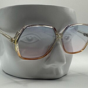 May include: Vintage oversized sunglasses with a clear frame and gradient lenses. The lenses transition from blue to pink, with a gold-toned trim. The glasses have a geometric shape and are displayed on a white bust.