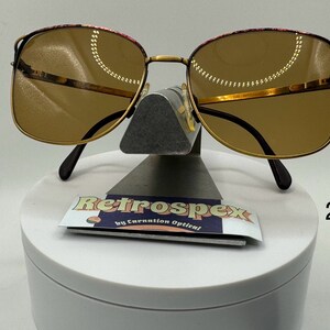 May include: Gold-tone square-shaped sunglasses with brown lenses and a burgundy-colored frame. The sunglasses are displayed on a white stand with a sticker that reads "Retrospecx by Carnation Optical" and the number "210".