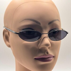 May include: Oval-shaped sunglasses with dark lenses and a thin, silver-colored frame. The sunglasses are displayed on a mannequin head. The lenses are a dark gray color, and the frame has a sleek, minimalist design.