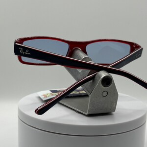 May include: Red and black Ray-Ban sunglasses with blue lenses, displayed on a silver stand.