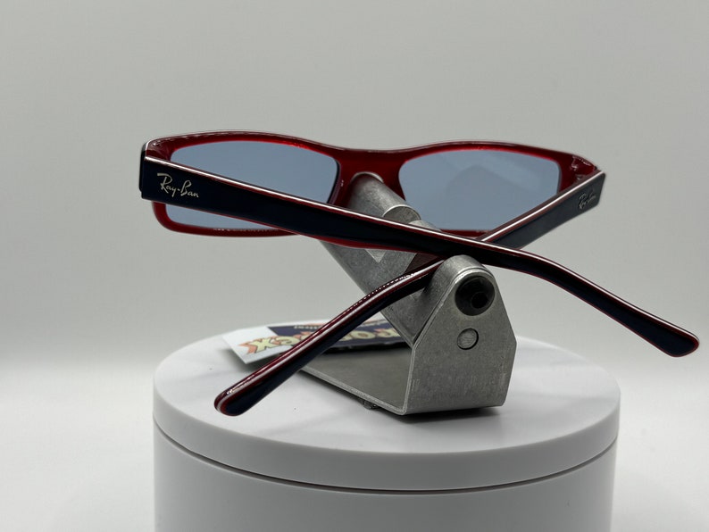 May include: Red and black Ray-Ban sunglasses with blue lenses, displayed on a silver stand.