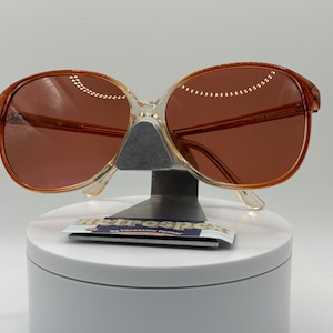 May include: A pair of clear, oval-shaped sunglasses with brown lenses. The sunglasses are on a white display stand with a small card that reads "Retrospec by Fashion Optica".