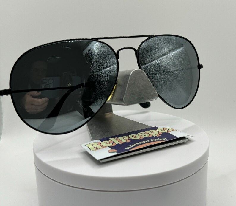 May include: Black aviator sunglasses with mirrored lenses. The sunglasses are on a white display stand with a business card that says "Retrospec by Salvation Optician".