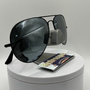 May include: Black aviator sunglasses with a silver metal nose bridge and dark gray lenses. The sunglasses are displayed on a white stand with a small card that reads "Retrospec by Independent Opticians".