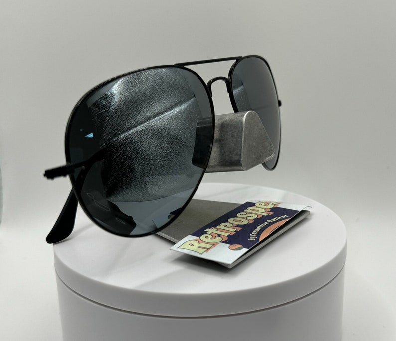 May include: Black aviator sunglasses with a silver metal nose bridge and dark gray lenses. The sunglasses are displayed on a white stand with a small card that reads "Retrospec by Independent Opticians".