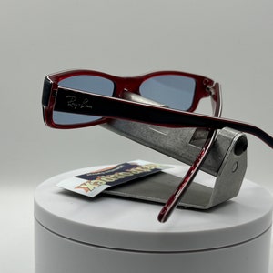 May include: Red Ray-Ban sunglasses with a black frame and blue lenses. The sunglasses are displayed on a silver stand with a white base.
