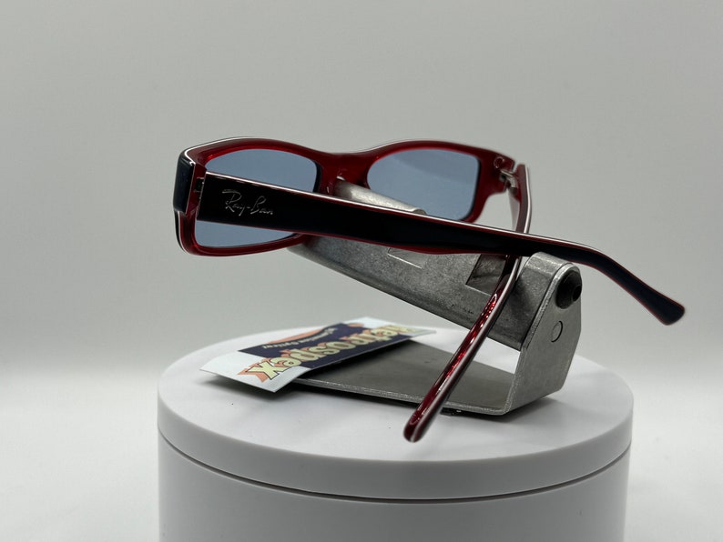 May include: Red Ray-Ban sunglasses with a black frame and blue lenses. The sunglasses are displayed on a silver stand with a white base.