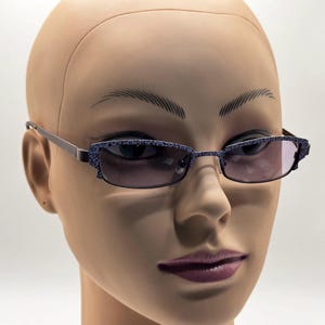 May include: Rectangular eyeglasses featuring a dark purple frame with a detailed black floral pattern on the front rims and temples. The lenses have a light purple gradient tint, and the thin metal arms connect to the frame with a simple hinge.