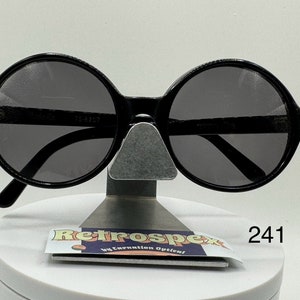 May include: Black round sunglasses with a black frame and dark gray lenses. The sunglasses are on a display stand with a white surface and a sticker that says "Retrospec by Carnation Optician" and "241".