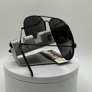 May include: Black aviator sunglasses with dark gray lenses, displayed on a silver stand. The sunglasses have a black frame and are resting on a white surface.