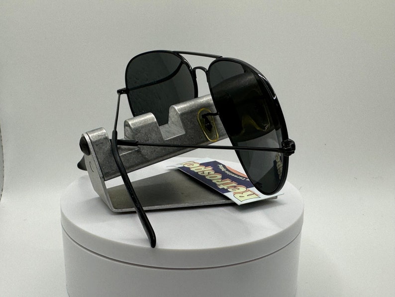 May include: Black aviator sunglasses with dark gray lenses, displayed on a silver stand. The sunglasses have a black frame and are resting on a white surface.