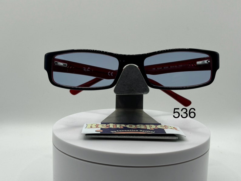 May include: Black rectangular eyeglasses with red frames and blue tinted lenses. The glasses are on a display stand with a white base and a black metal stand. The number 536 is printed on the base of the stand.