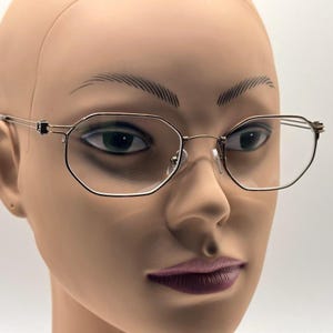 May include: Gold-toned, octagonal eyeglasses with clear lenses. The frames have a thin, delicate design with a bridge and nose pads. The glasses are displayed on a mannequin head, showcasing the eyewear's shape and style.