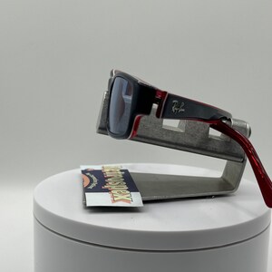 May include: Red and black sunglasses with a silver metal frame and the text "Ray-Ban" on the side. The sunglasses are on a silver metal stand.