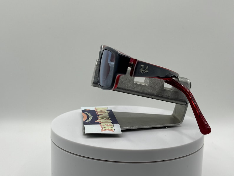 May include: Red and black sunglasses with a silver metal frame and the text "Ray-Ban" on the side. The sunglasses are on a silver metal stand.