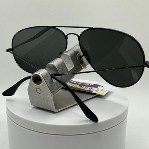 May include: Black aviator sunglasses with dark gray lenses. The sunglasses are on a silver stand with a white background.