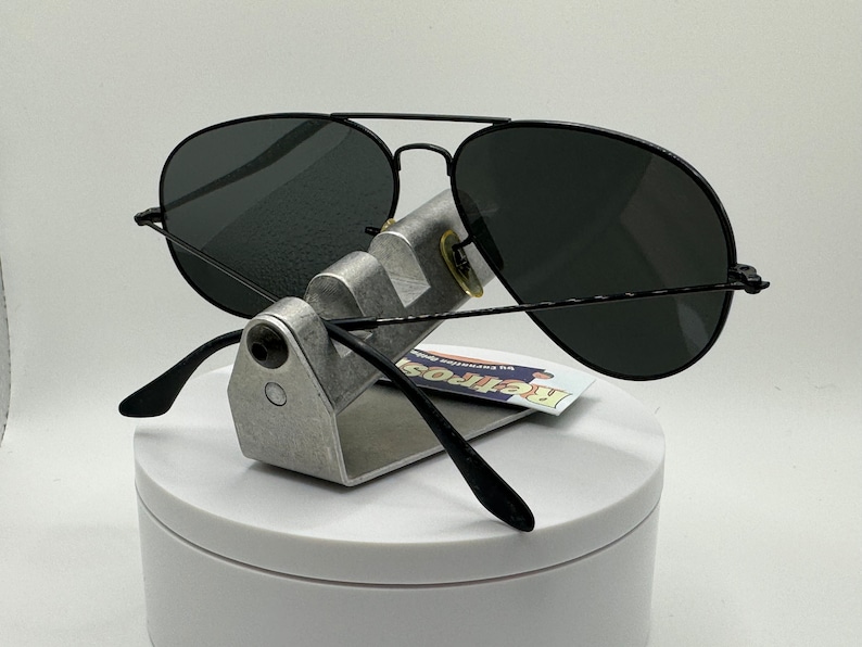May include: Black aviator sunglasses with dark gray lenses. The sunglasses are on a silver stand with a white background.