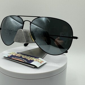 May include: Black aviator sunglasses with dark gray lenses. The sunglasses are on a white display stand with a sticker that says "Retrospecx by Coronation Optical".