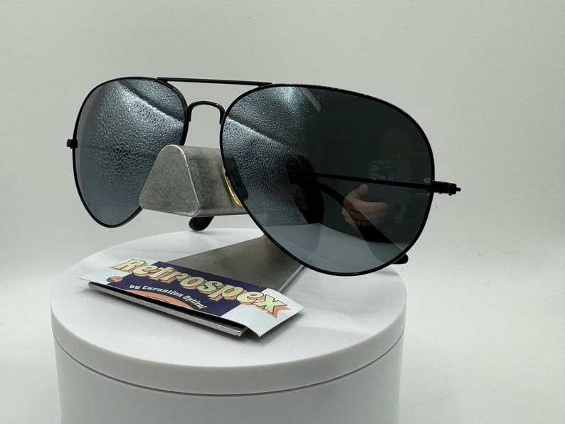 May include: Black aviator sunglasses with dark gray lenses. The sunglasses are on a white display stand with a sticker that says "Retrospecx by Coronation Optical".