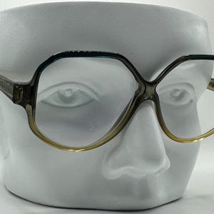 May include: Vintage eyeglasses with a geometric frame design. The frames are a gradient of colors, transitioning from a dark grey to a light yellow at the bottom. The glasses are displayed on a white bust.