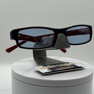May include: Black rectangular eyeglasses with red arms and blue tinted lenses. The glasses are displayed on a white stand with a small black and white label that reads "Retrospec" in a retro font.