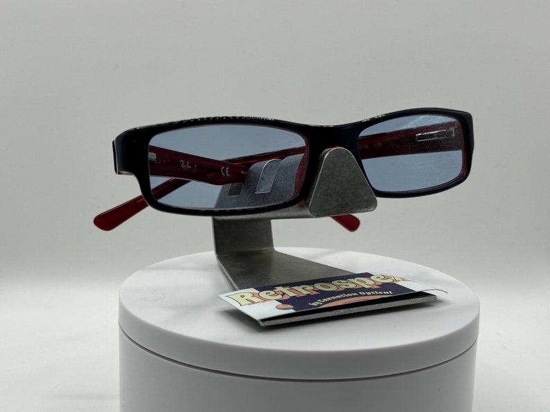 May include: Black rectangular eyeglasses with red arms and blue tinted lenses. The glasses are displayed on a white stand with a small black and white label that reads "Retrospec" in a retro font.