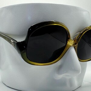 May include: Oversized sunglasses with a black and olive green frame and dark lenses. The sunglasses have a gold-colored trim around the lenses and a decorative element on the temples. The sunglasses are displayed on a white mannequin head.