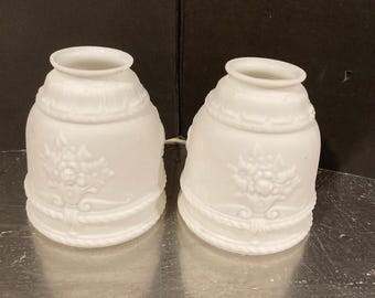 May include: A pair of white milk glass lamp shades with a bell shape. Each shade features a raised embossed floral bouquet design on the front, a rope-like texture around the base, and a smooth, flared opening at the top for light fixture attachment.
