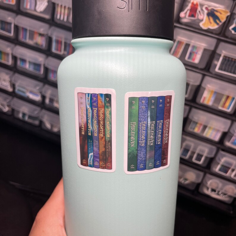 May include: A light blue water bottle with two stickers on it. The stickers feature the book covers for the Dragonwatch and Fablehaven series. The Dragonwatch sticker shows the covers for the first six books in the series, and the Fablehaven sticker shows the covers for the first five books in the series.