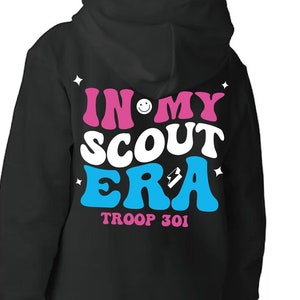 May include: Black hooded sweatshirt with a pink and blue graphic on the back that says "In My Scout Era" with a smiley face and a patch. The text "Troop 301" is below the graphic. The front of the sweatshirt has pink text that says "Ellie Troop 1234".