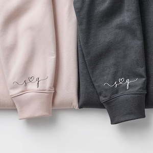 May include: Close-up of three sweatshirts in pink, charcoal gray, and light gray. Each sweatshirt sleeve cuff features a small embroidered design of a heart and script initials. The sweatshirts are folded and arranged side by side.