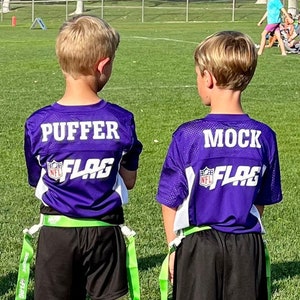 May include: Seven young athletes stand on a green field, wearing matching purple and white flag football jerseys. Each jersey has a name and the word "FLAG" printed on the back. They wear black shorts, green flags, and colorful socks.