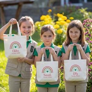 May include: Three off-white canvas tote bags, each featuring a printed rainbow graphic in pink, tan, and green, with a central heart. Below the rainbow, the names Chloe, Sarah, and Lily are printed in pink, followed by the text Troop 3364.