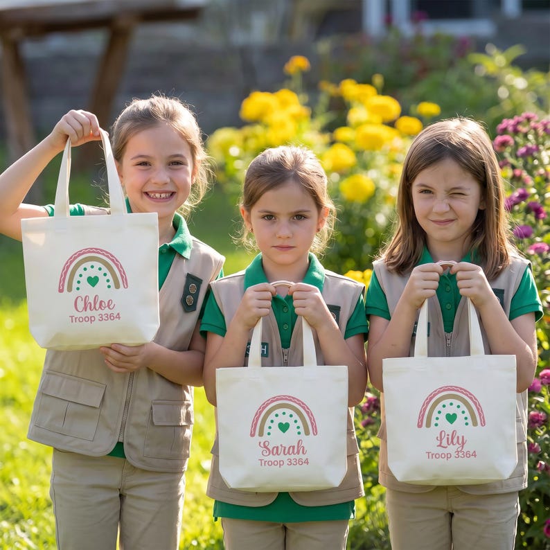 May include: Three off-white canvas tote bags, each featuring a printed rainbow graphic in pink, tan, and green, with a central heart. Below the rainbow, the names Chloe, Sarah, and Lily are printed in pink, followed by the text Troop 3364.