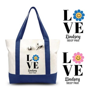 Pu&ograve; includere: Una borsa tote in tela bianca con rifiniture blu e una tasca con una grafica nera "LOVE" con una margherita blu e il testo "Lindsey TROOP 1468".