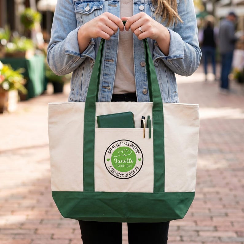 May include: A cream-colored canvas tote bag with dark green trim, handles, and a bottom panel. The front features a circular graphic with the text "Great Leaders Inspire Greatness In Others," "Janelle," and "Troop 1045," plus two pens and a green wallet.