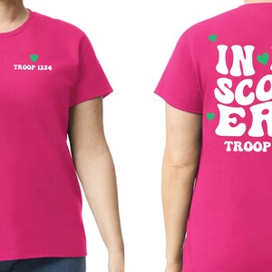 Może przedstawiać: R&oacute;żowa koszulka z białym sercem i tekstem "TROOP 1234" po lewej stronie. Z tyłu koszulki znajduje się napis "IN MY SCOUT ERA" w kolorze białym z zielonymi sercami i tekstem "TROOP 1234" poniżej.