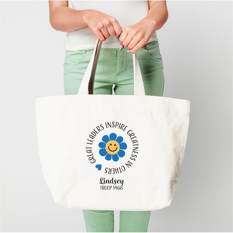 Pu&ograve; includere: Borsa tote in tela bianca con un fiore blu e un viso sorridente. Il testo intorno al fiore recita "Great Leaders Inspire Greatness in Others". Sotto il fiore c'&egrave; il testo "Lindsey Troop 1468".