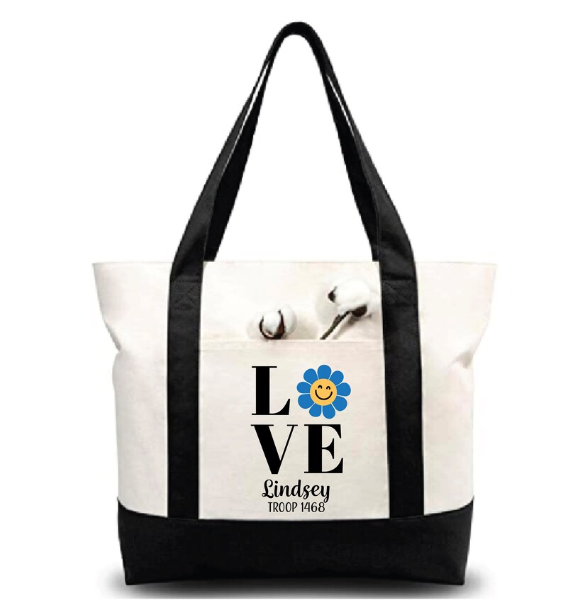 Pu&ograve; includere: Borsa tote in tela bianca con manici neri e fondo nero. La borsa ha una tasca sul davanti con un fiore nero e blu con un'emoticon gialla sorridente. Il testo "LOVE" &egrave; in lettere nere sopra il fiore e il testo "Lindsey TROOP 1468" &egrave; sotto il fiore.