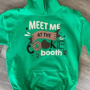 May include: Green hooded sweatshirt with the text "Meet me at the cookie booth" in white and brown lettering. A brown and white cookie graphic is included.